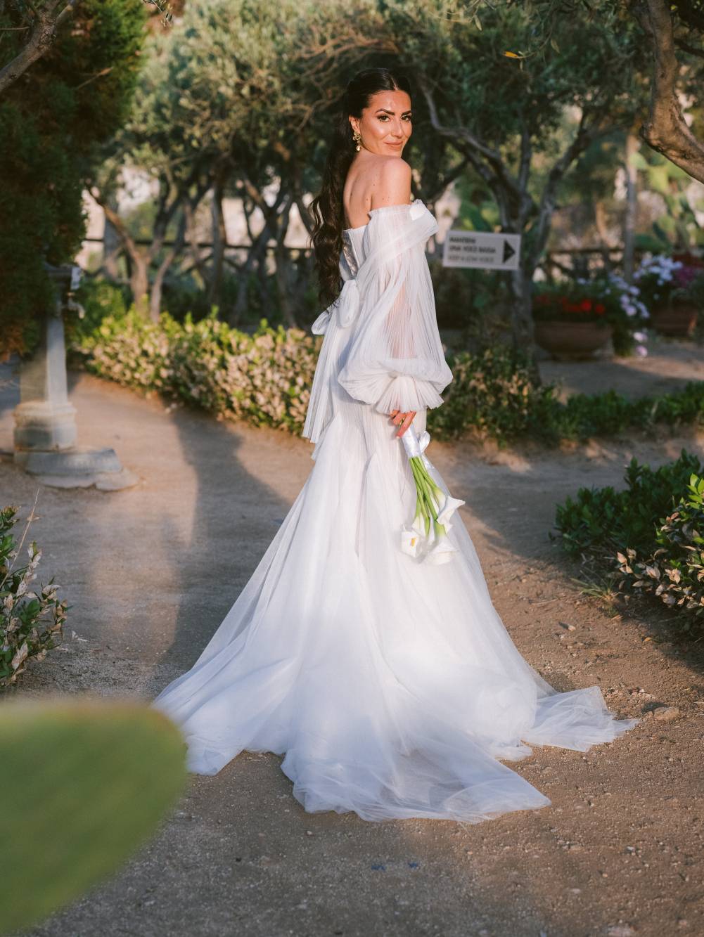 Marianna in piedi nel giardino della villa tra ulivi e arbusti fioriti, vista di profilo che si gira verso l'obiettivo, bouquet di calle