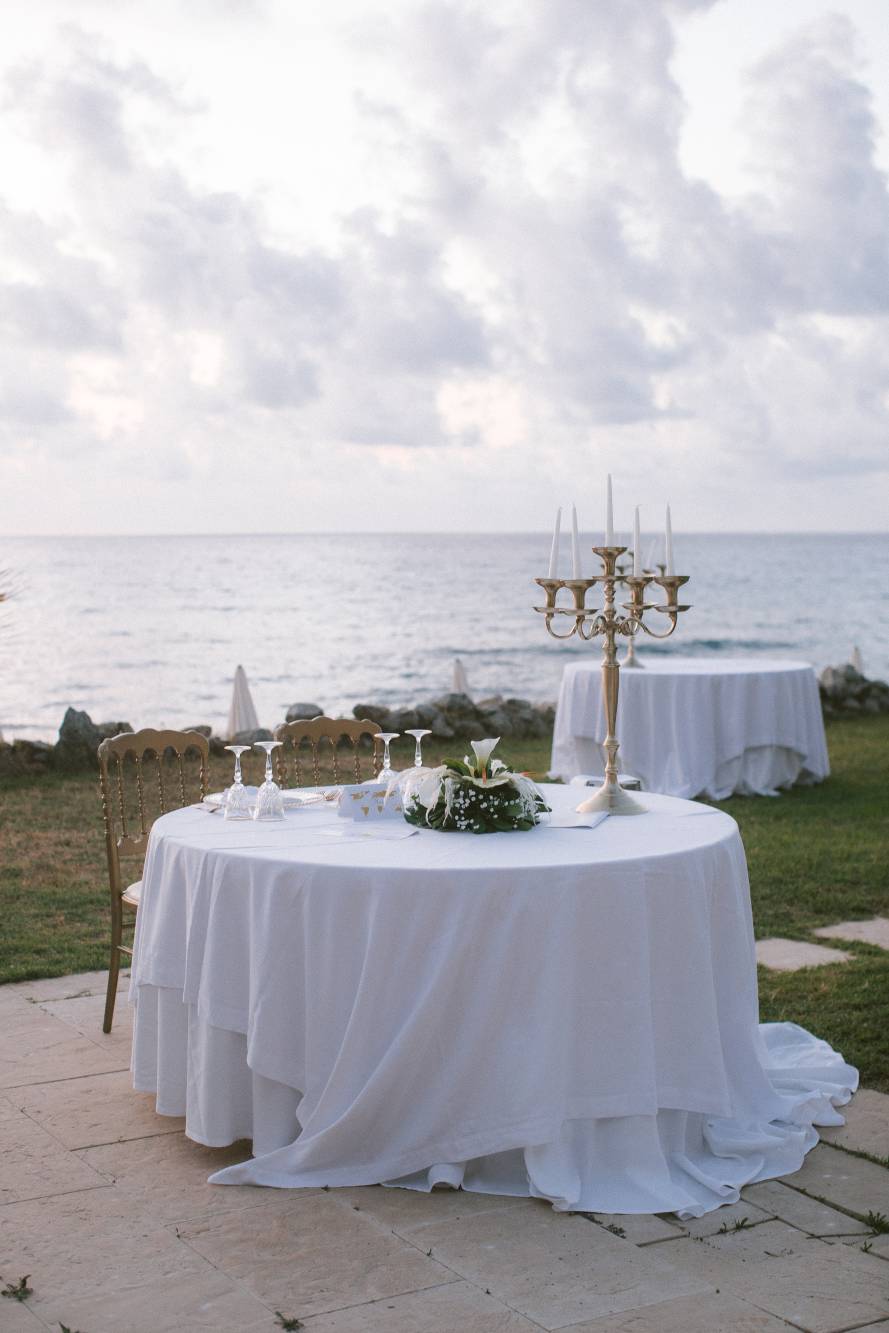 Tavolo degli sposi all'aperto con tovagliato bianco lungo, candelabro a 7 candele color ottone, composizione di calle bianche al centro, vista frontale sul mare al crepuscolo