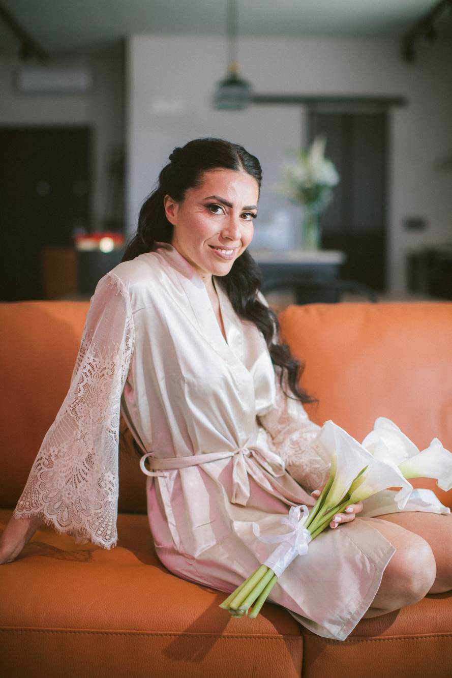 Marianna in vestaglia di seta tiene un bouquet di calle bianche, divano arancione e finestra alle spalle