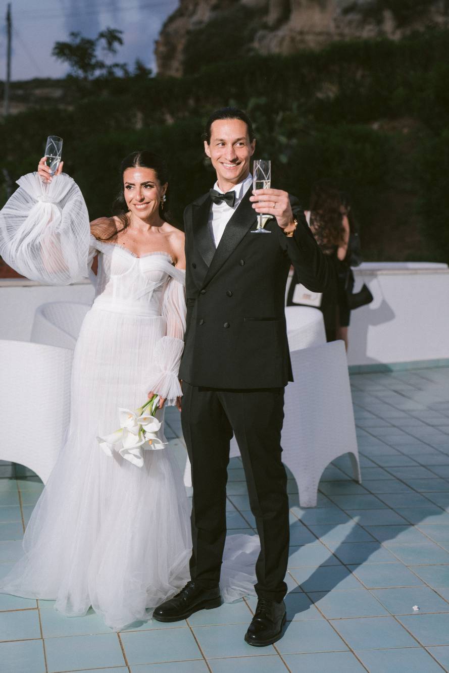 Marianna e Andrea brindano in piedi sul terrazzo del Sunshine, ora del crepuscolo, scogliera rocciosa alle spalle, pavimentazione color azzurro