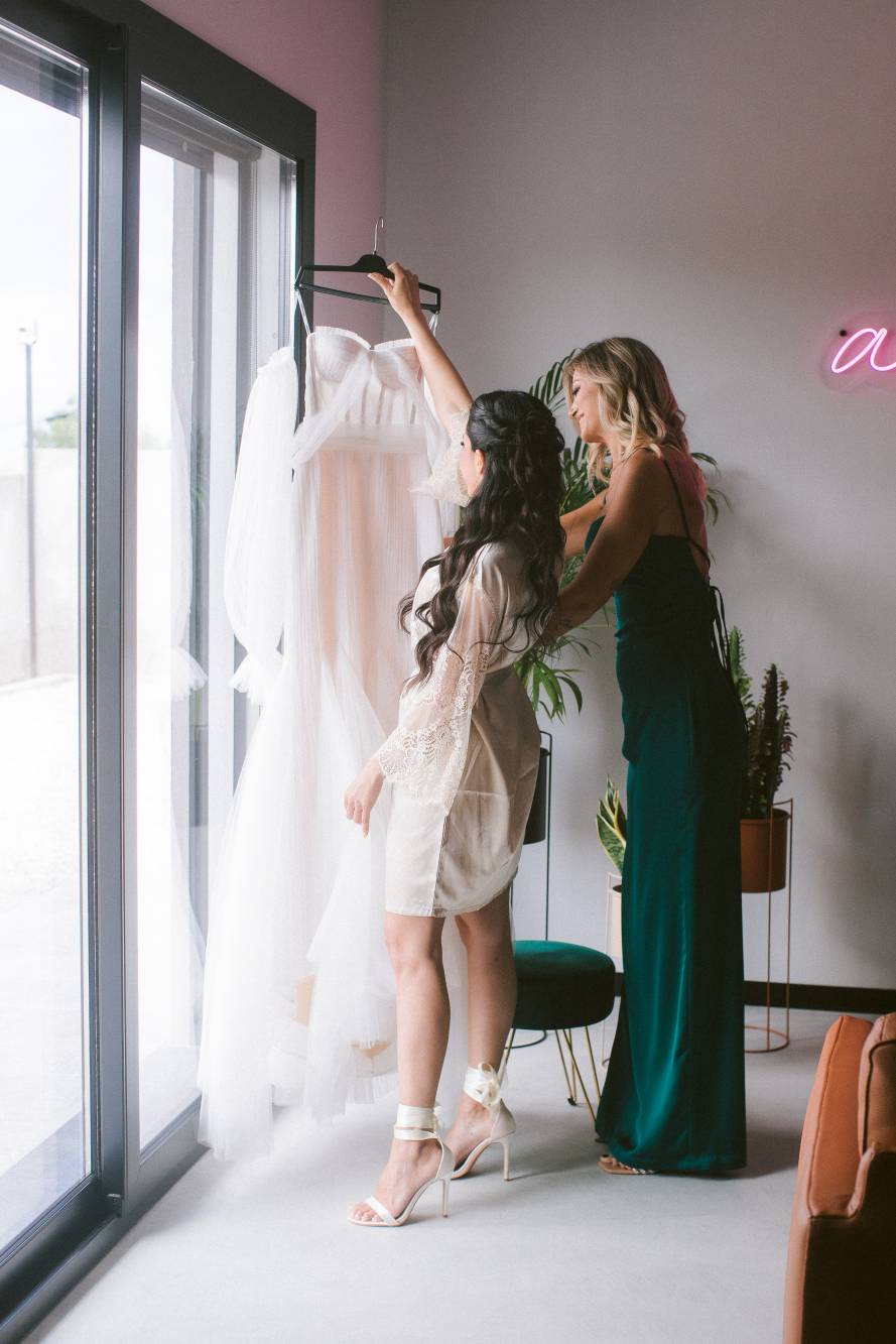 Marianna in vestaglia con damigella in abito verde smeraldo prendono il vestito appeso davanti alla finestra, insegna neon 'amore' sulla parete
