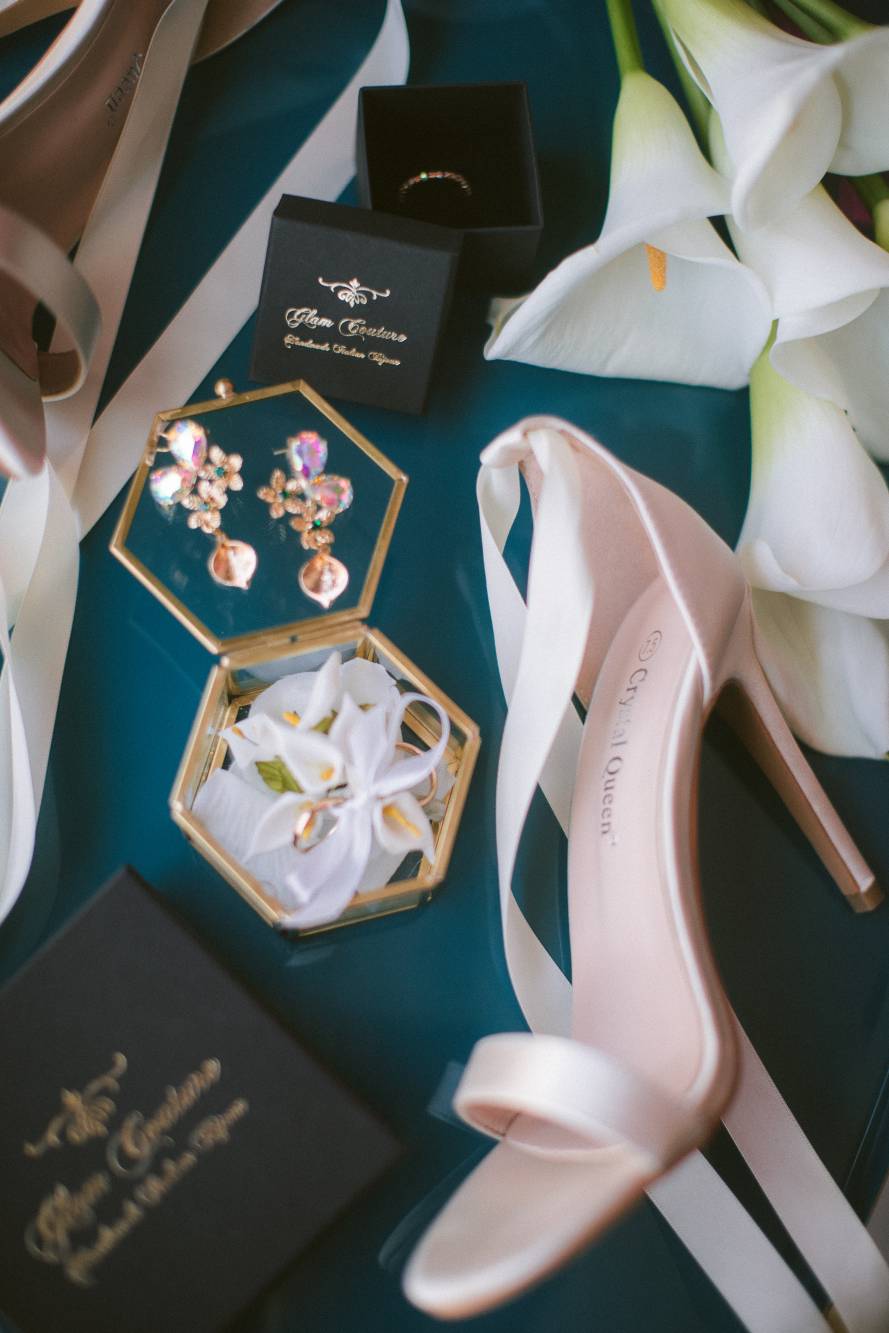 Dettagli getting ready: scarpe rosa Crystal Queen, scatola anelli Glam Couture aperta con calle, orecchini gemma multicolor su vassoio verde petrolio