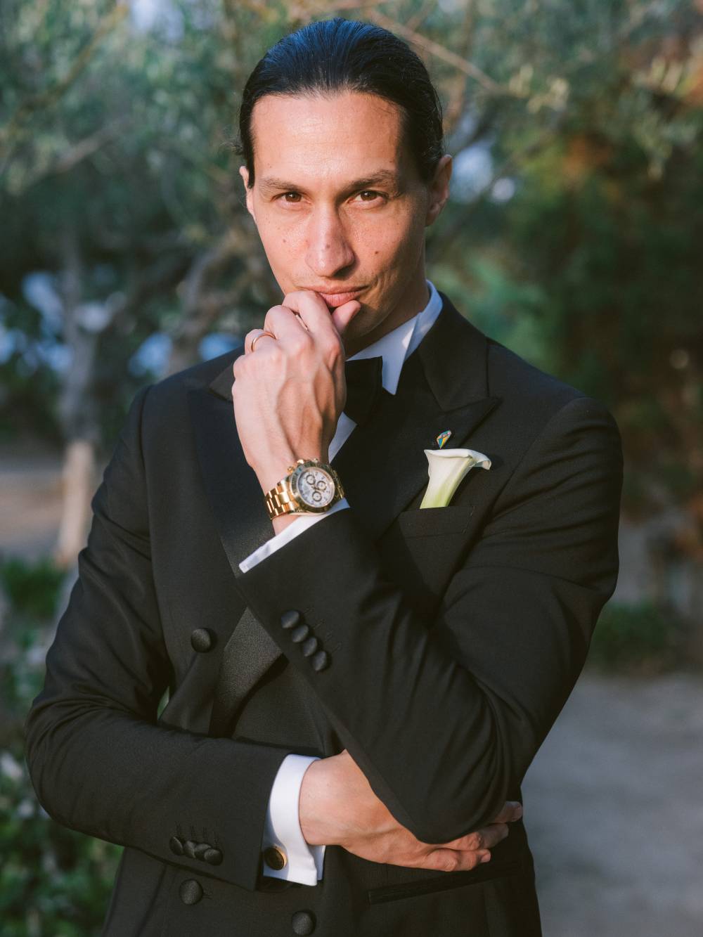 Ritratto dello sposo Andrea: primo piano, smoking doppiopetto, bouttoniere di calla bianca, Rolex Daytona oro, dietro ulivo